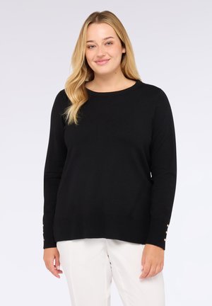Mujer sonriente con cabello rubio, vistiendo un suéter negro de manga larga con botones en los puños y pantalones blancos, de pie sobre un fondo claro.