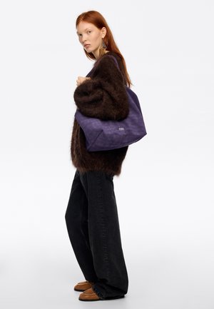 Purpurinės spalvos audinio peties krepšys su tekstūruota paviršiaus struktūra, laisvo stiliaus dizainas ir baltas logotipas. Modelis dėvi tamsų, pūkuotą megztinį ir juodas džinsus.