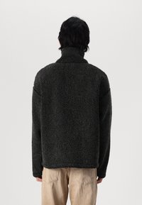 Chaqueta de forro polar negra con un alto cuello, que presenta una superficie texturizada y suave, con hombros caídos, combinada con pantalones en un tono beige claro.