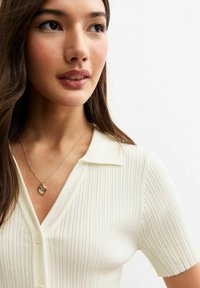 Geribbelde zachte crème trui met korte mouwen en een kraaghalslijn, met een delicate gouden hartketting en natuurlijke haartextuur.
