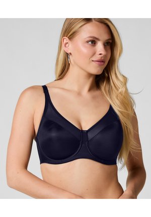 Blonde Frau trägt einen marineblauen Vollschalen-BH mit verstellbaren Trägern und posiert vor einem hellgrauen Hintergrund.