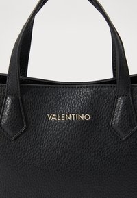 Black textured leather handbag featuring two short handles and a gold logo detail, with a structured shape and minimalist design.