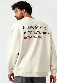 Felpa oversize color crema realizzata in tessuto morbido. Presenta scritte nere e rosse sul retro con un collo e polsini a costine.