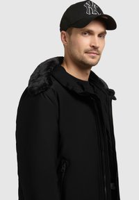 Melnā polsterētā jaka ar mākslīgās kažokādas apkakli, ar rāvējslēdzēja priekšu un sānu kabatām. Valkāta kopā ar melnu cepuri ar stilizētu logo.