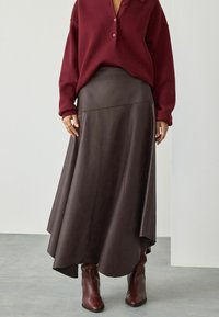 Donna che indossa un maglione bordeaux con bottoni, una gonna in pelle marrone scuro asimmetrica e stivali marroni a punta, in piedi su un pavimento grigio.