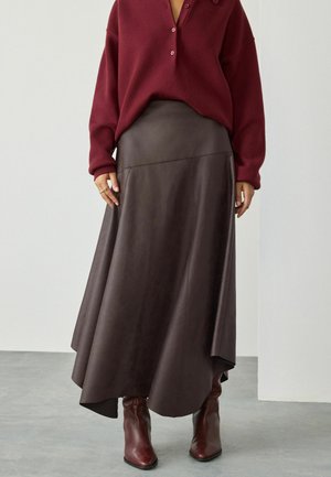 Femme portant un pull bordeaux à boutons, une jupe en cuir marron foncé asymétrique, et des bottes brunes à talons pointus, se tenant sur un sol gris.