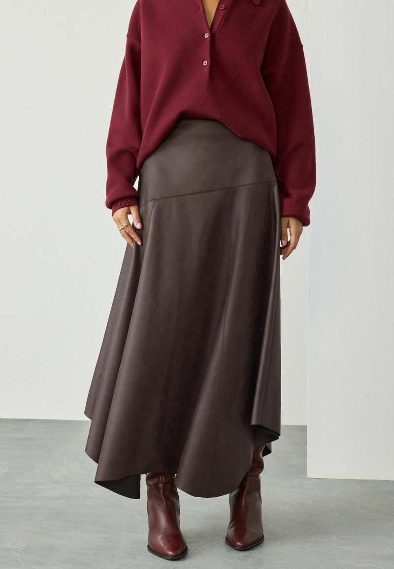 Donna che indossa un maglione bordeaux con bottoni, una gonna in pelle marrone scuro asimmetrica e stivali marroni a punta, in piedi su un pavimento grigio.