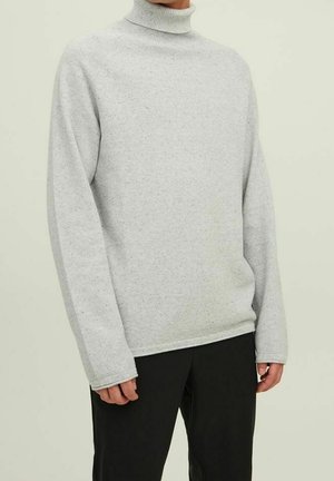 Personne portant un pull à col roulé gris clair à manches longues et un pantalon noir, debout devant un fond clair uni.