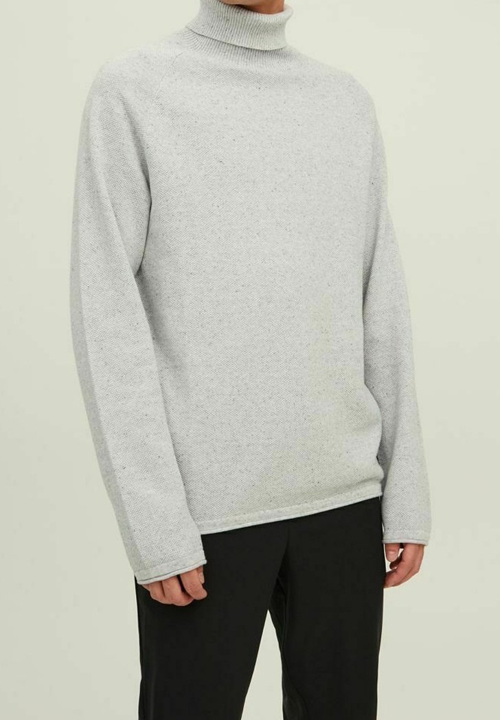 Personne portant un pull à col roulé gris clair à manches longues et un pantalon noir, debout devant un fond clair uni.