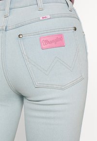 Jasnoniebieskie dżinsy z różowym naszywką Wrangler na tylnej kieszeni oraz małą metką Barbie umieszczoną nad kieszenią na pasek.