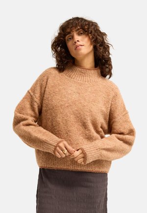 Donna con capelli ricci che indossa un maglione marrone chiaro e peloso e una gonna scura texturizzata, posando con le mani vicino alla vita su uno sfondo semplice.