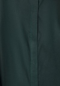 Tessuto verde scuro con una texture liscia, caratterizzato da una prominente cucitura verticale evidenziata da un filo azzurro.