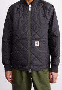 Sort quiltet jakke med lynlås foran, ribstrikkede manchetter og et Carhartt-logo påsat. Har to sidelommer og en glat tekstur.