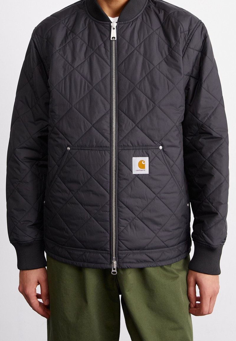 Sort quiltet jakke med lynlås foran, ribstrikkede manchetter og et Carhartt-logo påsat. Har to sidelommer og en glat tekstur.