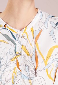 Blouse blanche ornée d'un motif floral abstrait en bleu, jaune et orange. Boutons sur le devant avec une texture douce et lisse.