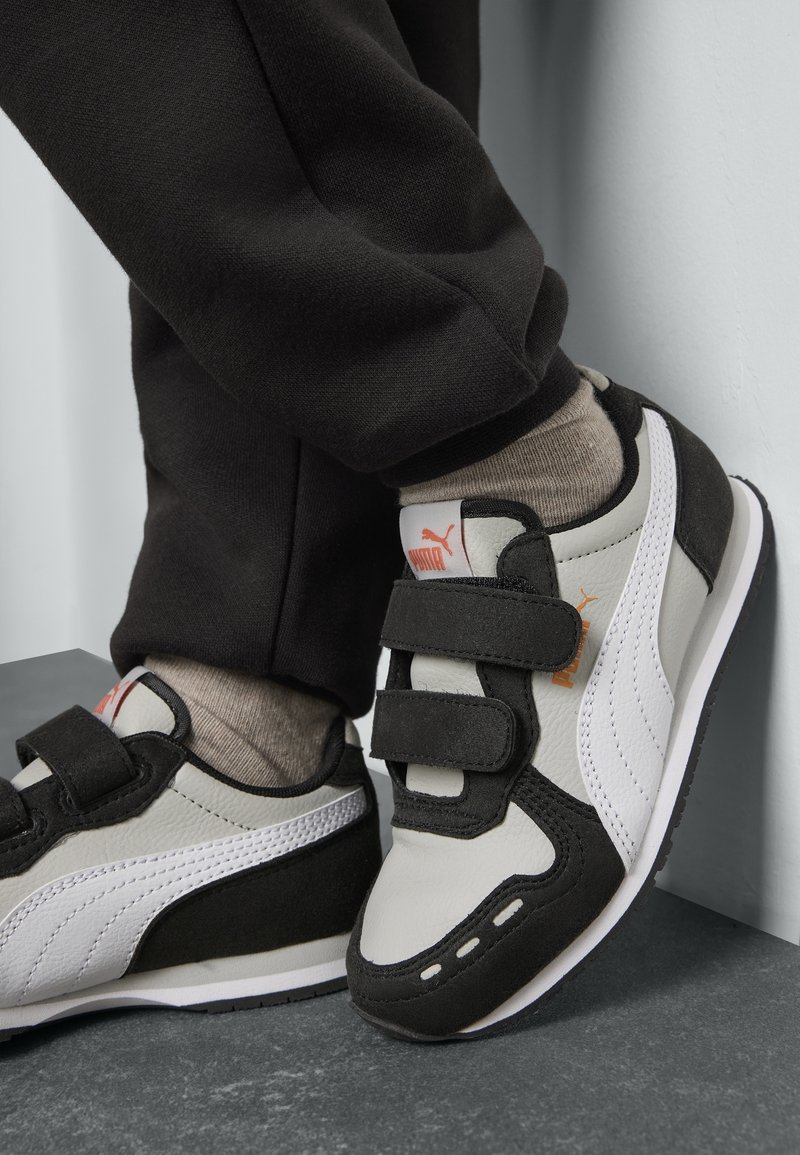 Enfant portant des baskets Puma noires et blanches avec des bandes Velcro, des chaussettes beige et un pantalon noir, debout sur un sol gris près d’un mur blanc.