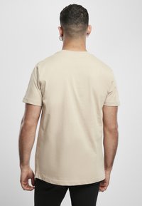 Muž s krátkými tmavými vlasy a náušnicemi, oblečený v jednoduchém béžovém tričku s krátkým rukávem, stojí zády k nám na světle šedém pozadí.