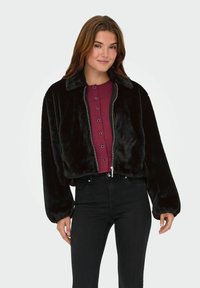 Zwart nepbont korte jas met een kraag, rits aan de voorkant en brede mouwen, gecombineerd met een bordeaux ribbeltop en zwarte jeans.