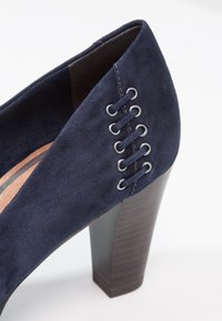 Námorní modré semišové boty na vysokém podpatku s blokovým podpatkem a ozdobnými stříbrnými oky provlečenými na boku. Hladký povrch a zaoblený design špičky.