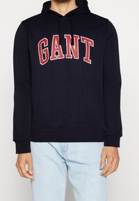 Sweat à capuche bleu marine avec un large logo "GANT" rouge en forme incurvée. Confectionné en matière douce avec une poche kangourou à l'avant et une capuche ajustable avec cordon de serrage.