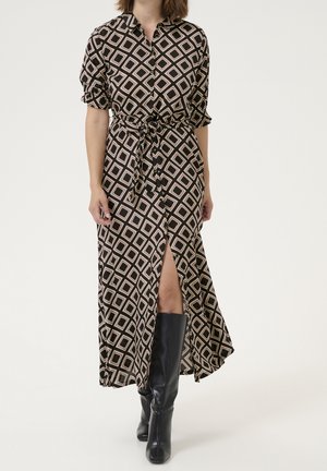 Femme portant une longue robe noire avec des carrés géométriques beiges, boutonnée devant, taille nouée, fente dévoilant la jambe, et bottes noires montantes.