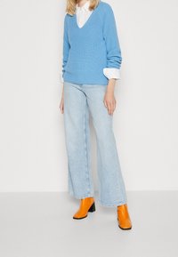 Maglione azzurro chiaro a righe con scollatura a V, camicia bianca con colletto sotto, abbinato a jeans chiari a zampa d'elefante e stivali arancioni con tacco.