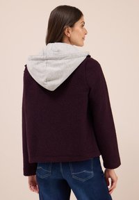 Veste en polaire bordeaux avec un sweat à capuche gris, présentant une surface texturée, une coupe droite et des manches longues. Portée avec un jeans en denim bleu.