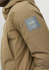 Kaki vattentät jacka med huva, som har ett texturerat tyg och en blå etikett på ärmen med koordinater och varumärkesnamn.