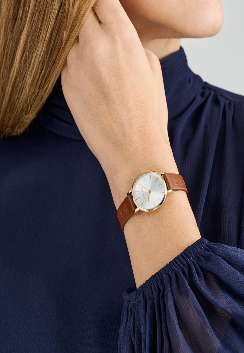 Okrągły zegarek z białą tarczą, złotą kopertą i brązowym skórzanym paskiem. Pasek ma wytłoczone litery. Noszony na nadgarstku o jasnym odcieniu skóry.