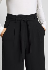 Pantalon noir taille haute avec une large ceinture et un détail de noeud à l'avant. Caractéristiques : poches latérales et plis marqués, fabriqué en tissu lisse.
