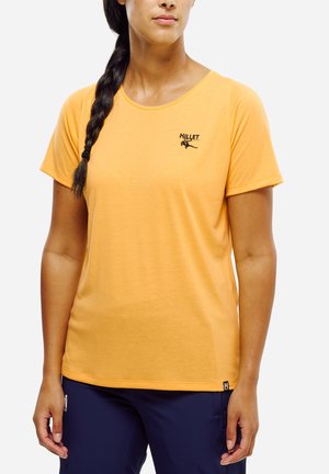 Femme portant un t-shirt orange Millet à manches courtes et un pantalon bleu foncé, avec une tresse sur l'épaule gauche, légèrement tournée vers la droite.