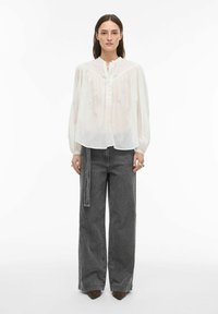 Valkoinen, puoliläpinäkyvä pusero, jossa on röyhelökaulus, edestä napillinen suljenta ja pitkät hihat, yhdistetty korkeavyötäröisiin, leveälahkeisiin harmaisiin farkkuihin.