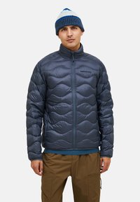 Blaues Pufferjacke mit gestepptem Muster, Reißverschluss vorne, hohem Kragen und schwarzem Logodetail. Kombiniert mit braunen Hosen und einer blauen Mütze.