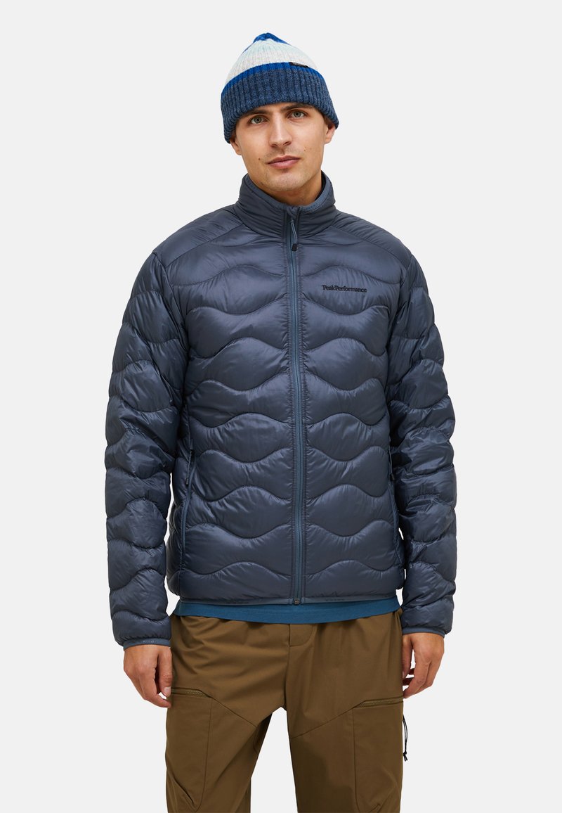 Blaues Pufferjacke mit gestepptem Muster, Reißverschluss vorne, hohem Kragen und schwarzem Logodetail. Kombiniert mit braunen Hosen und einer blauen Mütze.