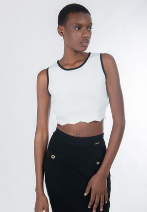Vrouw met kort haar, gekleed in een wit cropped mouwloos topje en een zwarte rok, kijkt opzij met een neutrale gezichtsuitdrukking.