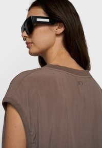 Mujer con cabello largo y castaño, que lleva gafas de sol negras de gran tamaño y una camiseta de manga corta color topo con el número "10" en el cuello trasero.