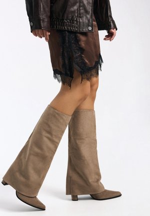 Bruine leren jas, bruine satijnen rok met zwarte kanten rand, lichtbruine suède kniehoge laarzen met spitse neus en blokhak.