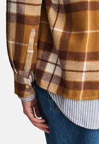 Camisa de franela a cuadros marrón y amarilla con puño de manga abotonado. Colocada sobre una camisa de rayas azul y blanca. Corte casual, textura suave.