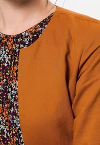 Primer plano de una mujer que lleva una chaqueta color naranja quemado sobre una blusa negra plisada con flores naranjas, blancas y amarillas.