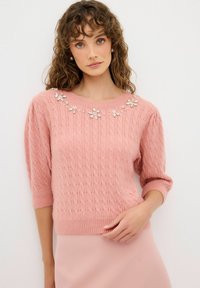 Pull en laine rose avec un motif en câble texturé, orné d'accents floraux embellis au niveau du col et de manches trois-quarts.
