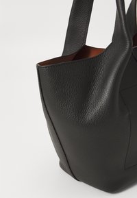 Black leather tote bag with a textured surface, open top, and slender handles. Interior shows brown lining. Angular shape with stitched details.