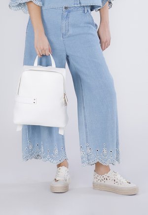 Persona che indossa jeans larghi ricamati azzurri e sneaker bianche con plateau e tagli, tenendo uno zaino in pelle bianca per la maniglia.