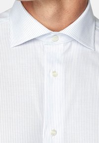 Vit skjorta med ljusblå vertikala ränder, knäppning med krage och två framtill knappar. Tyget verkar slätt med en fin struktur.