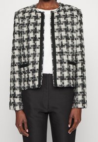 Personne portant une veste à carreaux noir et blanc texturée sur un haut blanc et un pantalon noir, debout les bras détendus le long du corps.