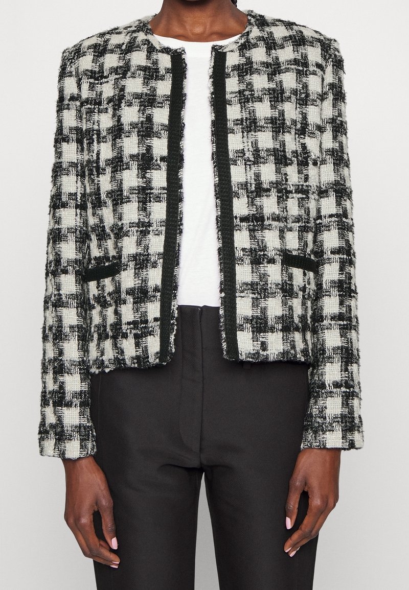 Personne portant une veste à carreaux noir et blanc texturée sur un haut blanc et un pantalon noir, debout les bras détendus le long du corps.