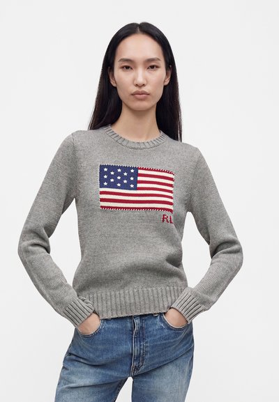 Femme aux longs cheveux foncés portant un pull gris en maille avec un motif drapeau américain et un jean bleu, mains dans les poches, expression neutre.