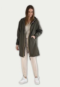 Imperméable vert olive avec une fermeture éclair et une capuche, agrémenté d'accents jaunes. Associ é à un pantalon beige et des baskets blanches. Texture lisse.