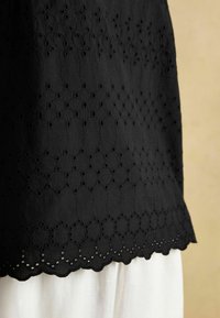 Melna auduma ar sarežģītu izšuvumu un izgrieztu rakstu, ar viļņotu apakšmali. Uzlikta virs baltas drēbes.