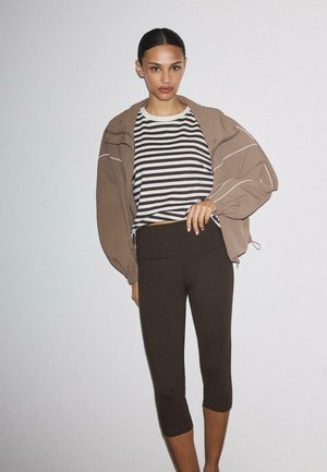 Kobieta z gładko zaczesanymi włosami, ubrana w pasiastą koszulę, brązowe krótkie legginsy i luźną brązową kurtkę na jasnym, jednolitym tle.