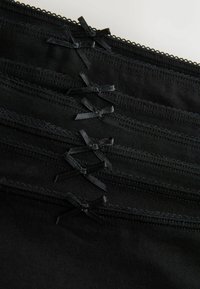 Stoh päť čiernych pásikov z látky na spodnú bielizeň s malými čiernymi ozdobnými mašličkami a vlnitými okrajmi, zobrazených zblízka.